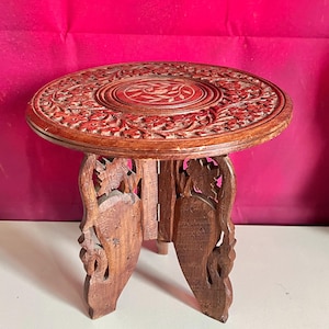 May include: A small, round, wooden side table with intricate floral carvings on the tabletop and legs. The table is a warm brown color and has a three-legged base. The tabletop is approximately 12 inches in diameter.