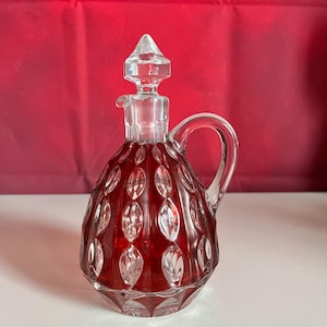 May include: A clear glass cruet with a ruby red interior and a clear glass stopper. The cruet has a handle and a diamond-cut pattern. The stopper is a faceted, pointed shape. The cruet is likely for serving oil or vinegar.