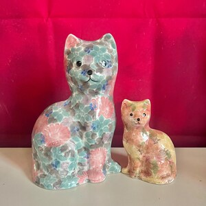 May include: Two ceramic cat ornaments. The larger cat is teal with pink and blue floral patterns. The smaller cat is orange, yellow, and pink floral patterns. Both cats are in a seated position.