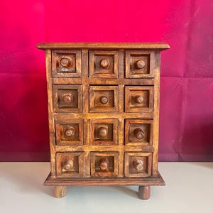 May include: A small, wooden cabinet with twelve square drawers. The cabinet is made of a warm-toned wood and has a simple, rustic design. Each drawer has a small, round knob. The cabinet sits on four short legs.