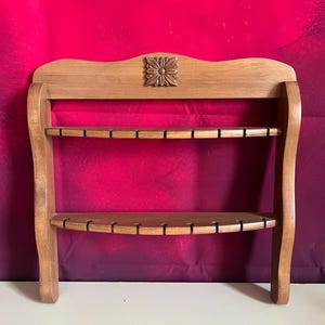Può includere: Un portacucchiaio in legno con due ripiani curvi, ciascuno con fessure per utensili. Il portacucchiaio è realizzato in legno marrone chiaro e presenta un'intaglio floreale decorativo nella parte superiore. Lo sfondo è rosso scuro.