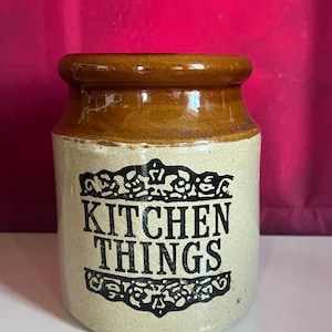 May include: A ceramic kitchen storage jar with a tan rim and base, and a cream-colored body. The words "KITCHEN THINGS" are printed in black, stylized lettering on the front of the jar.