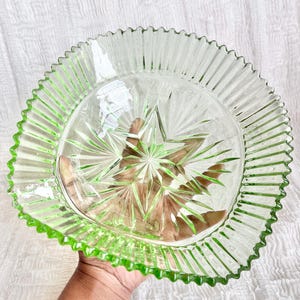Könnte beinhalten: Ein klarer Glasteller mit Sternmuster in der Mitte und einem gerüschten, hellgrünen Rand. Der Teller wird von einer Hand gehalten, die seine komplizierten Details und die lebendige Farbe hervorhebt. Ideal zum Servieren oder Dekorieren.