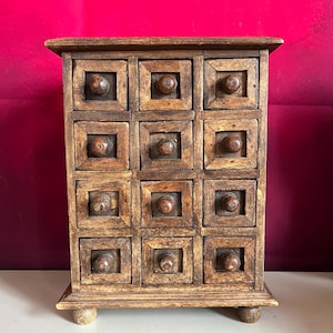 May include: A small, wooden apothecary cabinet with twelve square drawers. Each drawer has a round knob. The cabinet is made of light brown wood and sits on four small, rounded feet. The background is a deep red.