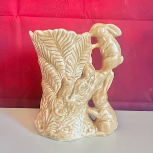 May include: A beige ceramic vase with a textured leaf design and three rabbit figurines climbing the side. The vase has a flared top and is set against a red background.