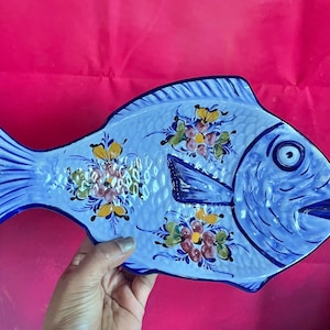 May include: A light blue ceramic fish-shaped serving platter with hand-painted floral designs in yellow, pink, and green. The platter has a dark blue outline and details, and is held against a red background.