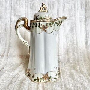 May include: A white porcelain teapot with a gold and green floral design. The teapot has a lid with a gold finial and a spout on the side.