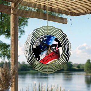 May include: A decorative metal wind spinner with a layered, wavy design. The center features a patriotic design with the word "VETERAN" on a red banner, a silhouette of a soldier, and a depiction of a weapon.