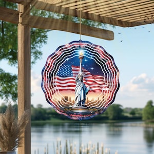 May include: A round, layered wind spinner featuring the Statue of Liberty, the American flag, and a starry night sky. The design incorporates red, white, and blue colors, with a wavy, layered edge. It hangs from a wooden structure, with a lake and trees in the background.