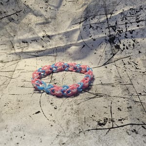 May include: A handmade bracelet made of small, interlocking rubber bands in shades of pink and light blue. The bracelet is circular and rests on a textured, off-white surface. The rubber band bracelet is a fun accessory.