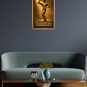 Könnte beinhalten: Gerahmtes Poster mit einer Vintage-Illustration eines Bodybuilders. Der Sepia-Ton-Postertext lautet "THE GOLDEN ERA of BODYBUILDING". Es hängt an einer dunkelblauen Wand über einem Sofa und einem Couchtisch mit Uhr und Vase.