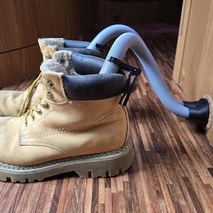 May include: A pair of tan work boots with yellow laces and a thick, gray sole. The boots are connected to a gray hose, likely for drying or ventilation. The boots are on a wooden surface.