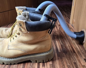 Könnte beinhalten: Ein Paar hellbraune Arbeitsstiefel mit gelben Schnürsenkeln und einer dicken, grauen Sohle. Die Stiefel sind mit einem grauen Schlauch verbunden, wahrscheinlich zum Trocknen oder zur Belüftung. Die Stiefel stehen auf einer Holzoberfläche.