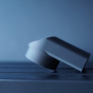 May include: A dark gray, angled object with a curved base, possibly a component or part of a larger item. The object is set against a gradient blue background, with a focus on its geometric shape and texture.