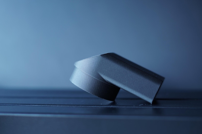 May include: A dark gray, angled object with a curved base, possibly a component or part of a larger item. The object is set against a gradient blue background, with a focus on its geometric shape and texture.