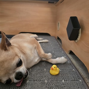 Könnte beinhalten: Ein hellbrauner Chihuahua-Hund liegt auf einer grauen Matte mit einem gelben Quietscheentchen. Der Hund liegt in der Nähe einer Holzkonstruktion mit einem schwarzen, geometrisch geformten Behälter.