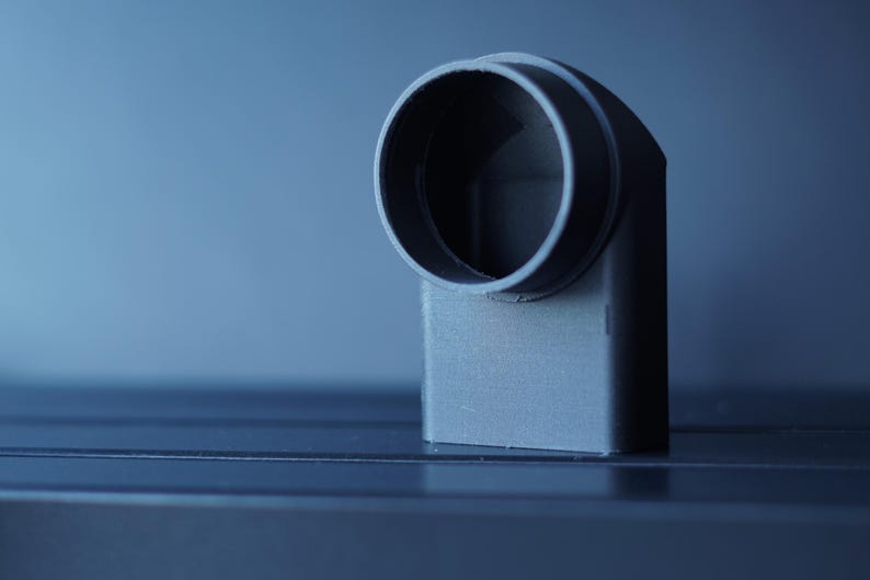 May include: A gray, right-angled pipe fitting with a circular opening. The fitting is made of a solid, matte material and is set against a gradient blue background. The design suggests a plumbing or industrial component.