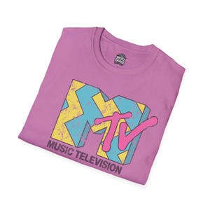 Könnte beinhalten: Ein lilafarbenes T-Shirt mit dem MTV-Logo im Retro-Design. Das Logo besteht aus blauen, gelben und rosa Elementen, darunter der Schriftzug "MUSIC TELEVISION". Das Shirt hat einen Rundhalsausschnitt und ein Etikett mit der Aufschrift "SHIRT SHACK CENTRAL".