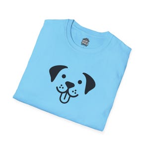 以下が含まれることがあります： 薄い青色のTシャツに、黒い漫画の犬の顔のグラフィックが描かれています。犬は舌を出しています。Tシャツの前には、「Shirt Shack Central」と書かれた小さなタグが付いています。