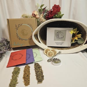 May include: An assortment of herbal products, including dried herbs, colorful packets, and a decorative box. A woven basket holds a card with floral and moon illustrations. The text on the card reads "Transforming the mind and spirit through nature's magic."