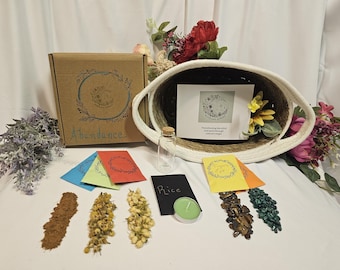 May include: An assortment of spiritual items, including a brown box labeled "Abundance", colorful cards, dried herbs, a small glass bottle, and a candle. A woven basket holds a card with text and flowers.