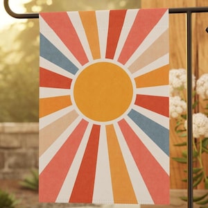 May include: A rectangular garden flag featuring a central yellow sun surrounded by radiating stripes in shades of orange, red, light pink, and muted blue, all separated by thin white lines against a white background.