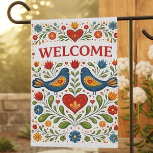 May include: A decorative garden flag with the word "WELCOME" in red. The flag features two blue birds with yellow hearts, surrounded by floral and heart designs in red, orange, and blue. The flag is displayed on a black metal stand.