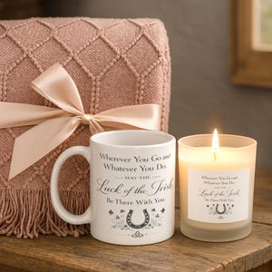 May include: A white ceramic mug and a frosted glass candle, both featuring the text "Wherever You Go and Whatever You Do. May the Luck of the Irish Be There With You." and a horseshoe graphic. A pink knit blanket with a satin ribbon is behind.