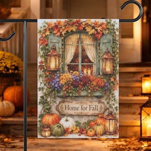 Puede incluir: Una bandera de jardín rectangular que muestra una ventana con contraventanas verdes, cortinas blancas y follaje otoñal. Una jardinera contiene flores coloridas. Debajo, un letrero dice Home for Fall, rodeado de calabazas y linternas.