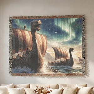 Op de afbeelding: Een geweven wandtapijtdeken met een scène van meerdere Viking-schepen met drakenkoppen als boeg die op onrustig water varen onder een nachtelijke hemel met noorderlicht. Het rechthoekige textiel heeft een gefranjerde rand en een gedetailleerd weefpatroon.