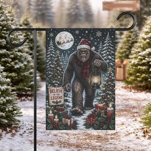 May include: A rectangular garden flag featuring a brown Bigfoot creature wearing a red and white Santa hat and a red and black plaid scarf, holding a lit lantern. The scene includes decorated Christmas trees, wrapped gifts, and a sign reading BELIEVE IN THE LEGEND under a full moon with a silhouette of a sleigh.
