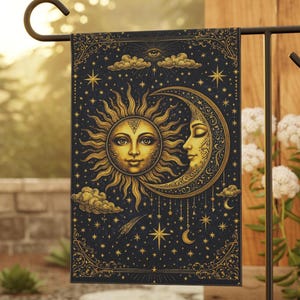 May include: A rectangular garden flag featuring a celestial design with a sun and moon face, stars, and clouds in gold against a dark blue background. The flag is hanging on a black metal stand, with white flowers in the background.