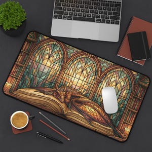 Könnte beinhalten: Eine rechteckige Schreibtischunterlage mit einer Buntglas-Illustration eines Drachen, der auf einem offenen Buch in einer Bibliothek schläft. Das Design zeigt Rundbogenfenster und Bücherregale mit dem Text ENTER THE REALM am oberen Bogen.