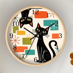 May include: A round wall clock with a light wood frame. The clock face features a black cat illustration, colorful geometric shapes, and black hands. The numbers are in a simple font.