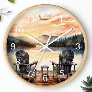 May include: Round wall clock with a light wood frame. The clock face features a watercolor painting of a lake scene with two Adirondack chairs, a small table, and a sunset. White clock hands and black numerals.