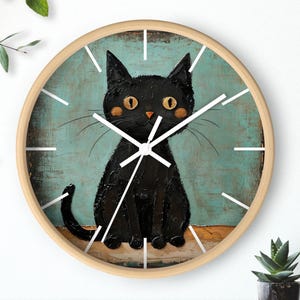 May include: A round wall clock with a light wood frame. The clock face features a black cat illustration with orange eyes and cheeks against a teal background. White clock hands and markers complete the design.