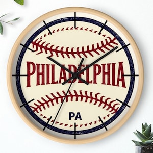 May include: Round wall clock with a baseball design. The clock face features the word "PHILADELPHIA" in red, with "PA" below. The clock has a light wood frame, black hands, and black hour markers.