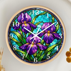 May include: Round wall clock with a light wood frame. The clock face features a stained glass design with purple iris flowers, green leaves, and a turquoise background. White numerals mark the hours.
