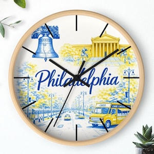 May include: Round wall clock with a light wood frame. The clock face features a Philadelphia city scene in blue and yellow, including the Liberty Bell, a museum, and a food truck. The word "Philadelphia" is written in script.