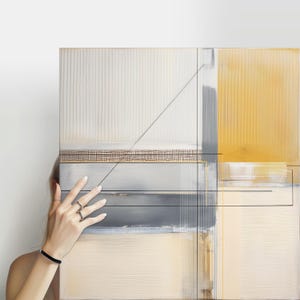 May include: Abstract art piece with a geometric design. The artwork features a combination of beige, gray, and yellow hues, with linear elements and a textured grid pattern. The piece is held by a person.
