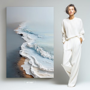 May include: A rectangular wall art print measuring 40 inches by 60 inches, featuring an aerial view of ocean waves crashing onto a sandy beach with a misty horizon. Beside the print stands a person wearing a white long-sleeve sweater and trousers.