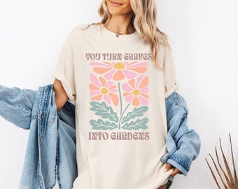 Può includere: T-shirt color crema con un design floreale rosa, pesca e verde-azzurro. La maglietta presenta la scritta "YOU TURN GRAVES INTO GARDENS" in un font retrò. Una giacca di jeans è drappeggiata sulle braccia.