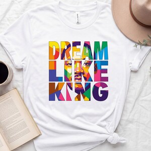 Può includere: T-shirt bianca con la scritta "DREAM LIKE KING" a blocchi colorati. Il design include un ritratto vibrante e stilizzato. Una tazza di caffè, un libro aperto e un cappello sono anche nell'immagine.