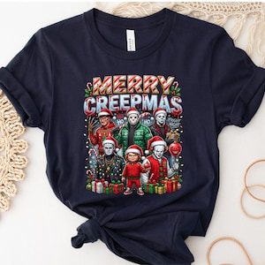 Puede incluir: Camiseta azul marino con un gr&aacute;fico festivo de personajes de pel&iacute;culas de terror con gorros de Pap&aacute; Noel, sosteniendo art&iacute;culos navide&ntilde;os. El texto "MERRY CREEPMAS" se muestra encima de los personajes. Tambi&eacute;n se incluyen regalos y bastones de caramelo.