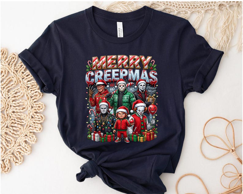 Puede incluir: Camiseta azul marino con un gr&aacute;fico festivo de personajes de pel&iacute;culas de terror con gorros de Pap&aacute; Noel, sosteniendo art&iacute;culos navide&ntilde;os. El texto "MERRY CREEPMAS" se muestra encima de los personajes. Tambi&eacute;n se incluyen regalos y bastones de caramelo.