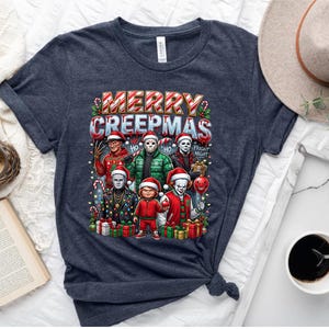 Puede incluir: Camiseta azul oscuro con el texto "MERRY CREEPMAS" e ilustraciones de personajes de pel&iacute;culas de terror con gorros de Pap&aacute; Noel. El dise&ntilde;o incluye bastones de caramelo, luces navide&ntilde;as y regalos. La camiseta es de un material suave.