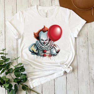 Puede incluir: Camiseta blanca con un gr&aacute;fico de un payaso aterrador sosteniendo un globo rojo. El payaso tiene pelo naranja, pintura facial blanca y un cuello con volantes. El dise&ntilde;o tiene un efecto de acuarela.