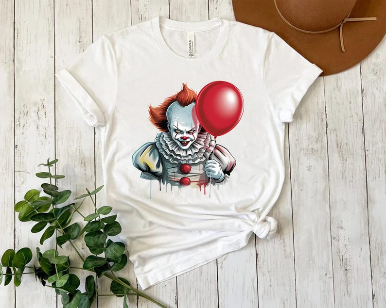 Puede incluir: Camiseta blanca con un gr&aacute;fico de un payaso aterrador sosteniendo un globo rojo. El payaso tiene pelo naranja, pintura facial blanca y un cuello con volantes. El dise&ntilde;o tiene un efecto de acuarela.