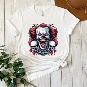 Puede incluir: Camiseta blanca con un dise&ntilde;o de payaso aterrador con pelo rojo, cara blanca y una sonrisa amenazante. El dise&ntilde;o incluye dos calaveras y globos rojos, con murci&eacute;lagos en el fondo. Un dise&ntilde;o espeluznante para Halloween.