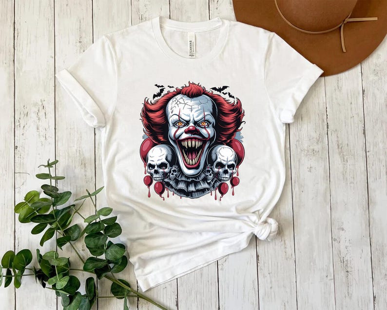 Puede incluir: Camiseta blanca con un dise&ntilde;o de payaso aterrador con pelo rojo, cara blanca y una sonrisa amenazante. El dise&ntilde;o incluye dos calaveras y globos rojos, con murci&eacute;lagos en el fondo. Un dise&ntilde;o espeluznante para Halloween.